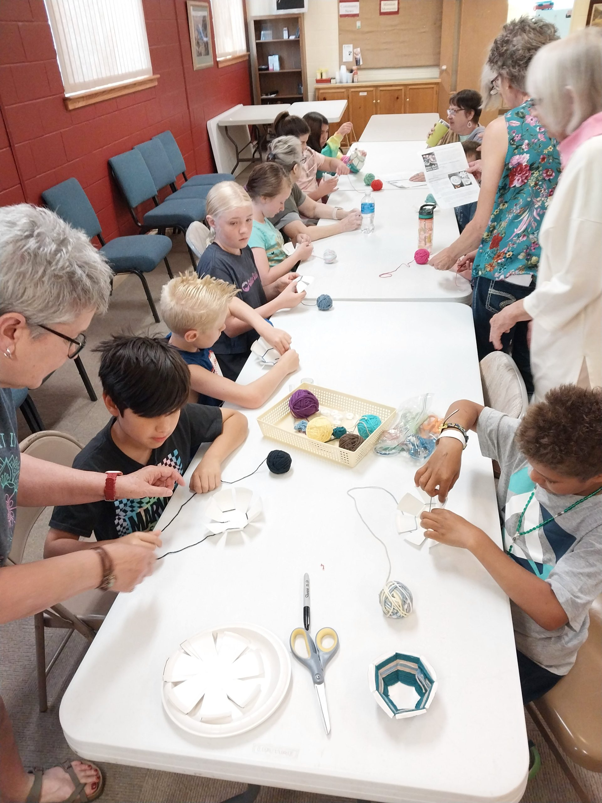 Group of children and adults crafting at a table.  They are making yarn decorations in a brightly lit room.