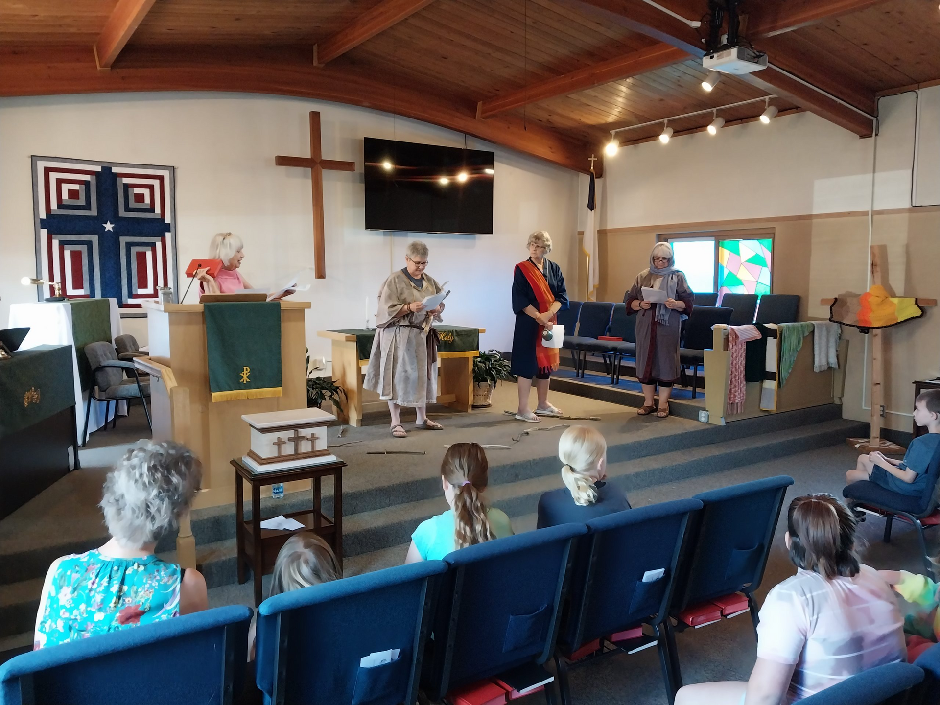 People in a church service; three women stand at podiums, reading. Audience in pews. Cross and artwork on walls.