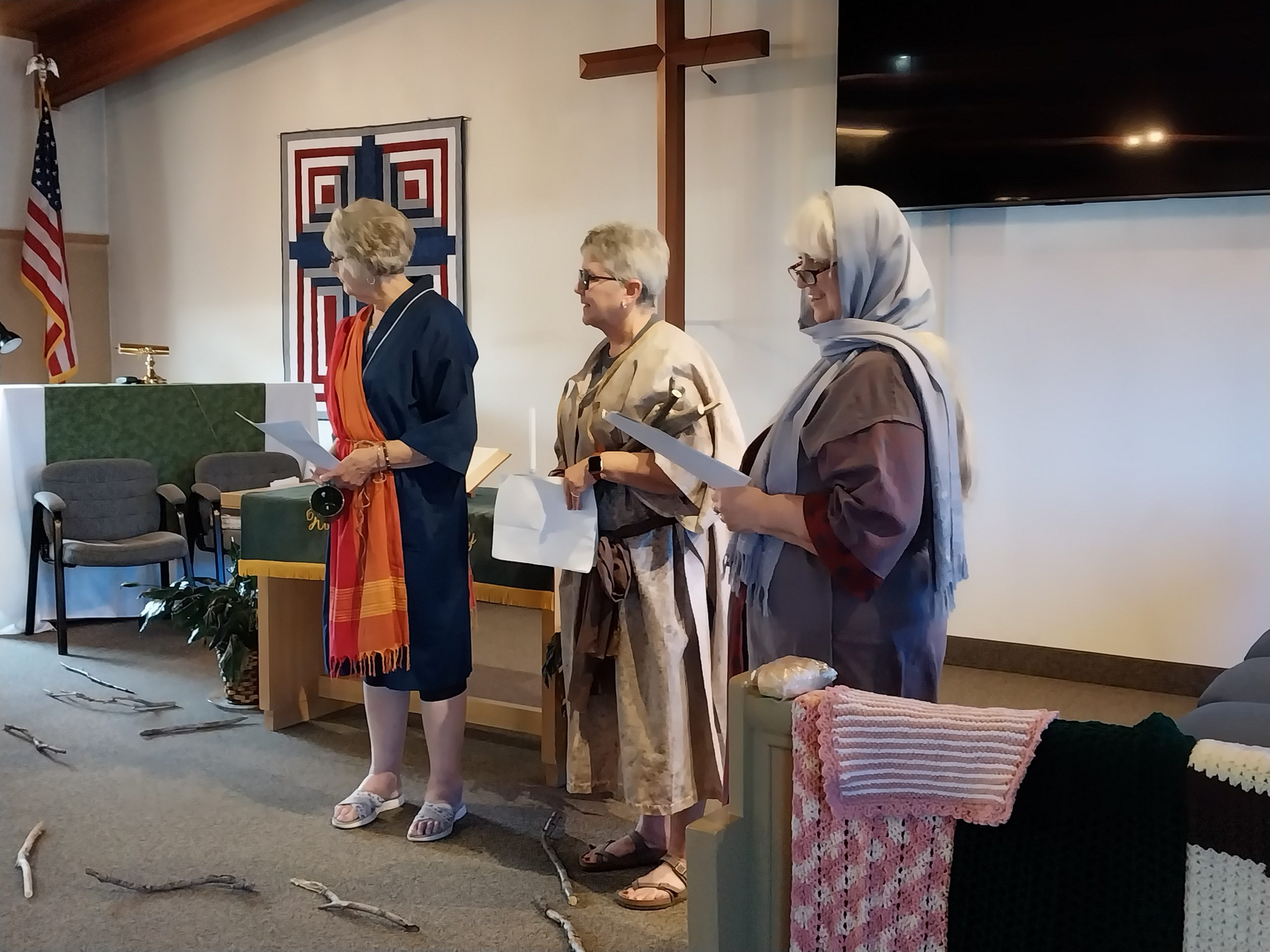 Three women in a church, possibly performing a play. One is draped in fabric, another in a robe. American flag visible.
