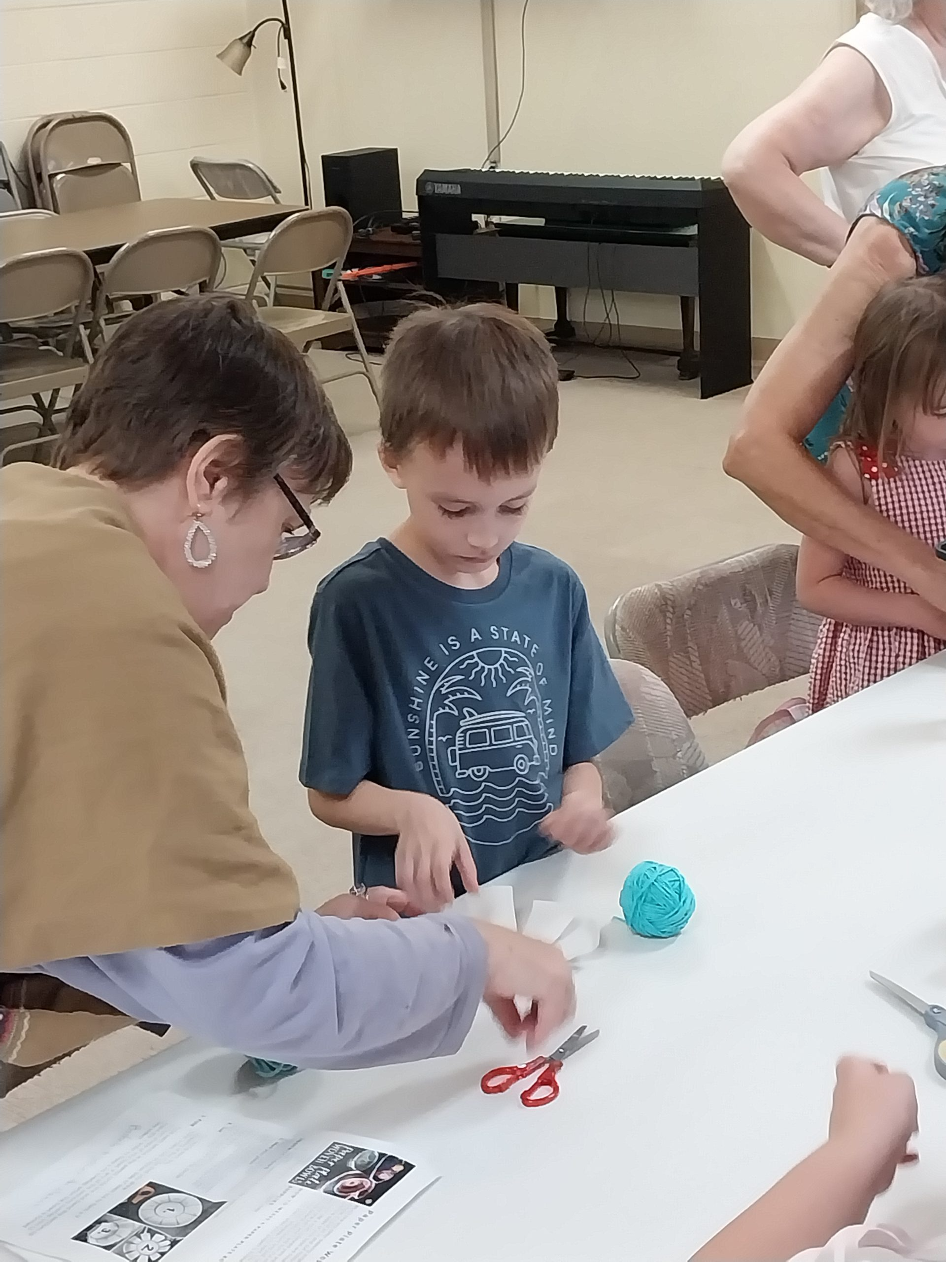 Woman and boy craft at a table, turquoise ball and red scissors present. Other people and a piano in the background.