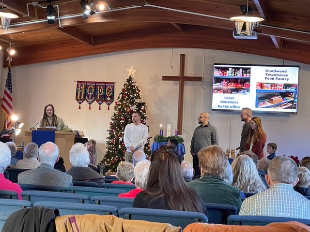 Church service with people, Christmas tree, cross, projected food pantry display, speaker.