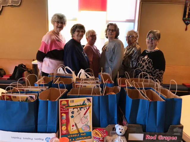 Six women stand behind blue gift bags on a table, likely for charity.