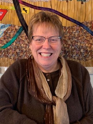 Woman in glasses smiles, wearing blue jacket and silver necklace against a gray background.