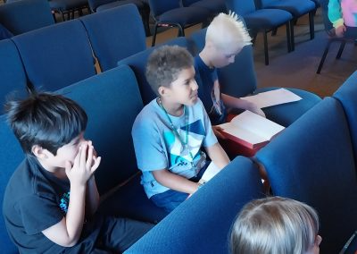 Three children seated in blue chairs in a room, two looking toward something, one with hands near face.