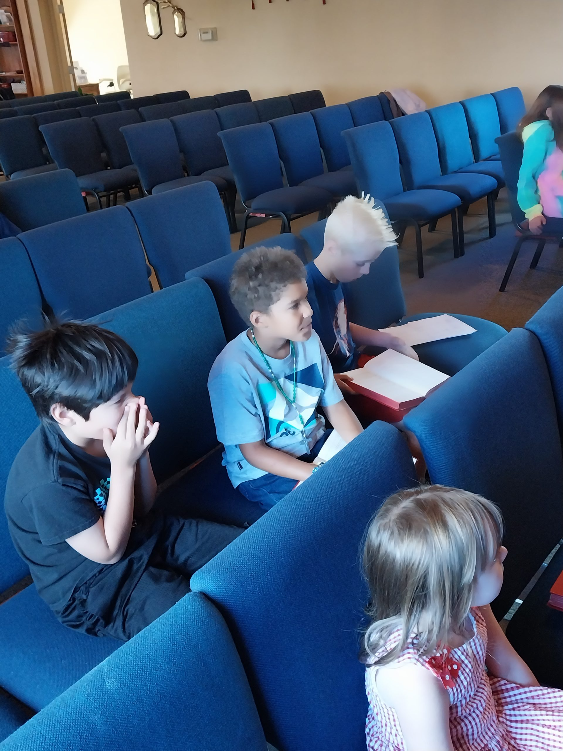 Children sitting in blue pews in a church. One boy is covering his mouth. Others have books.