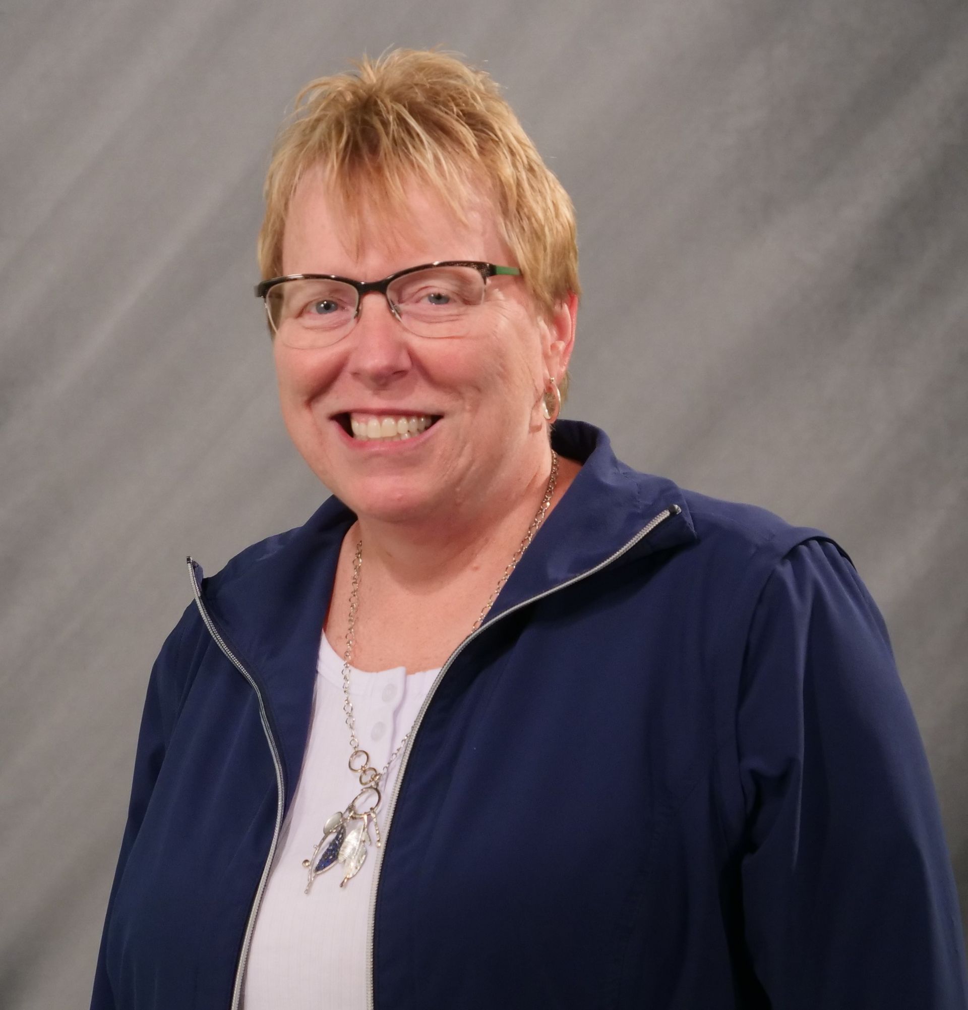 Woman in glasses smiles, wearing blue jacket and silver necklace against a gray background.