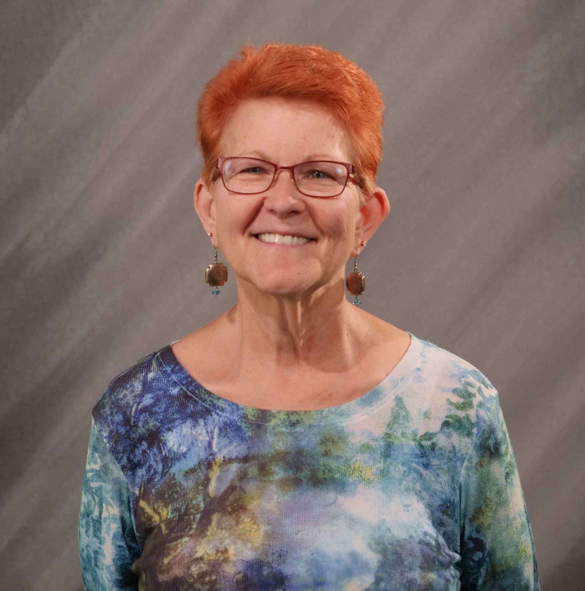 Woman with orange hair, glasses, smiling, wearing a blue and green patterned top, against a gray backdrop.