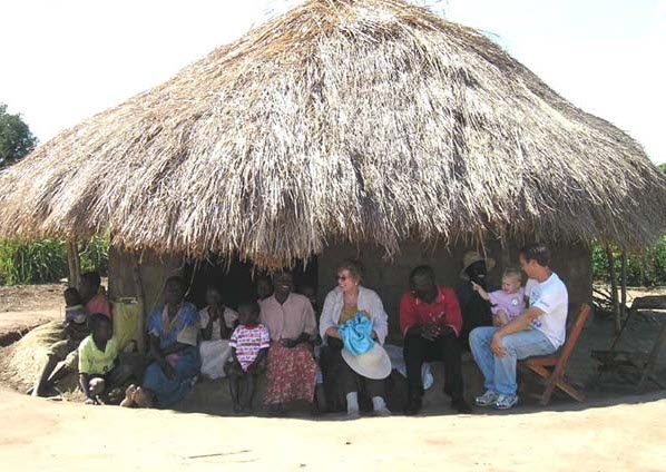 People sit under a thatched-roof hut. Some appear to be interacting, possibly in a village setting.