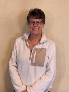 Woman in glasses smiles, wearing blue jacket and silver necklace against a gray background.