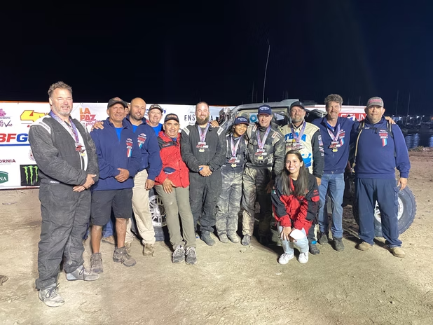 Group of people in racing suits posing together at night beside a race car.
