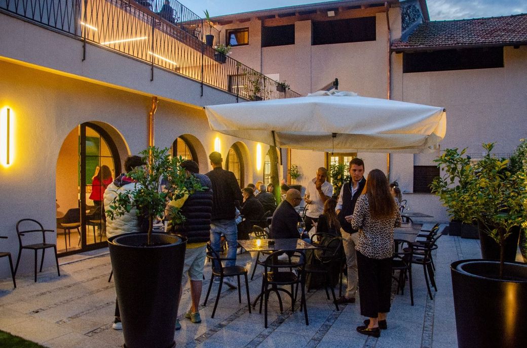 Un gruppo di persone è in piedi attorno a un patio con tavoli e sedie sotto un ombrellone.
