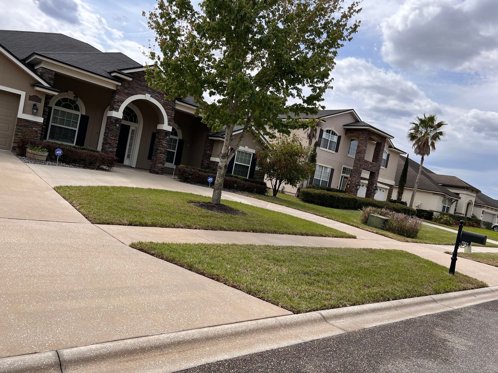 Suburban street with houses, green lawns, sidewalks, and a tree. Cloudy sky.