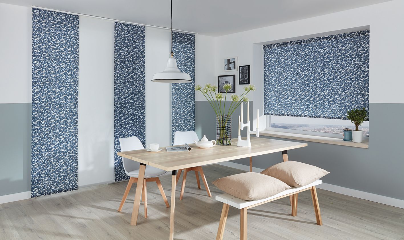 Dining room with a light wood table, bench, and chairs. Blue patterned panels and window shades, white walls.  Abundance Blinds In Charmhaven, NSW