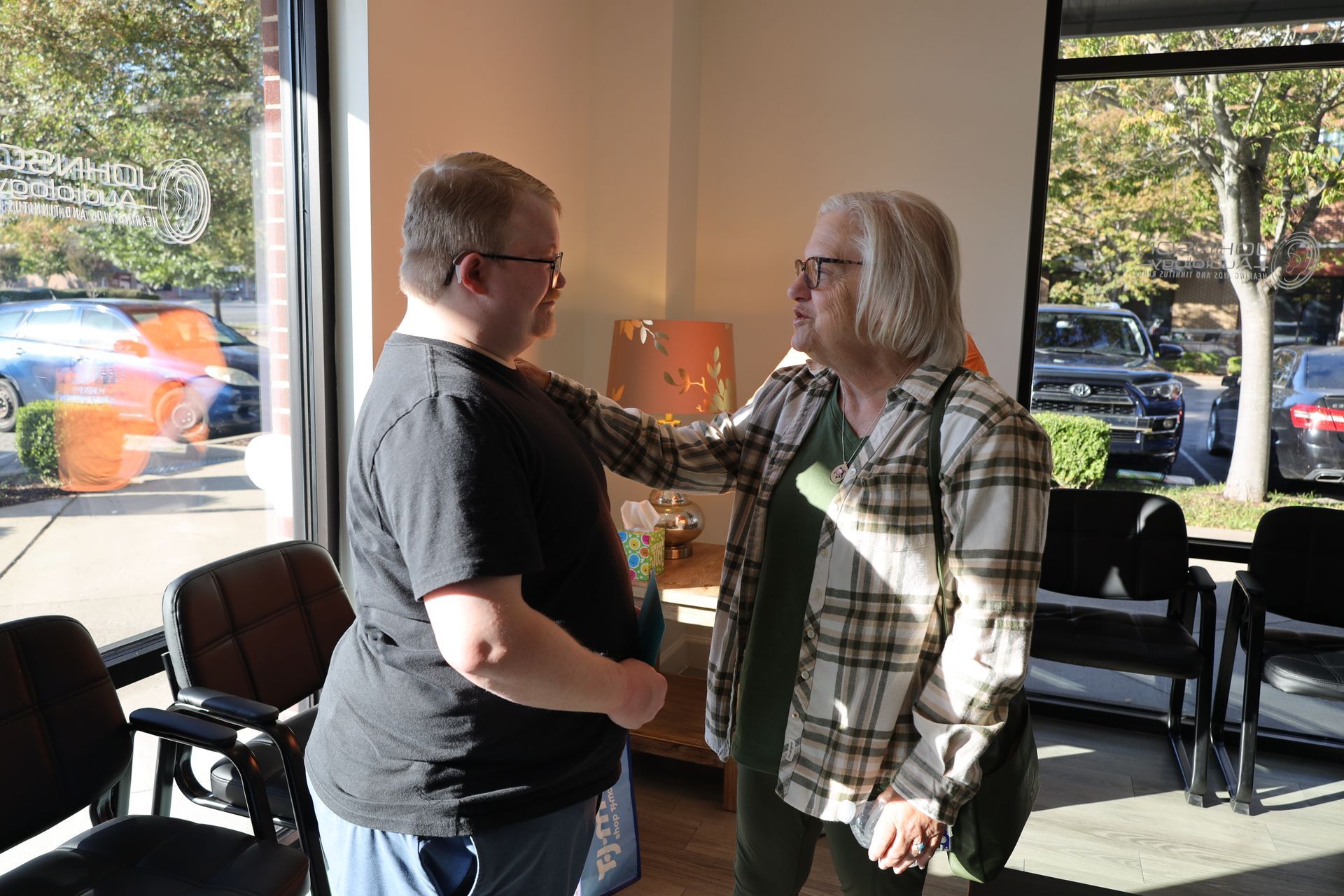 Two people talking near a window, one touching the other's arm. Sunlight streams in.