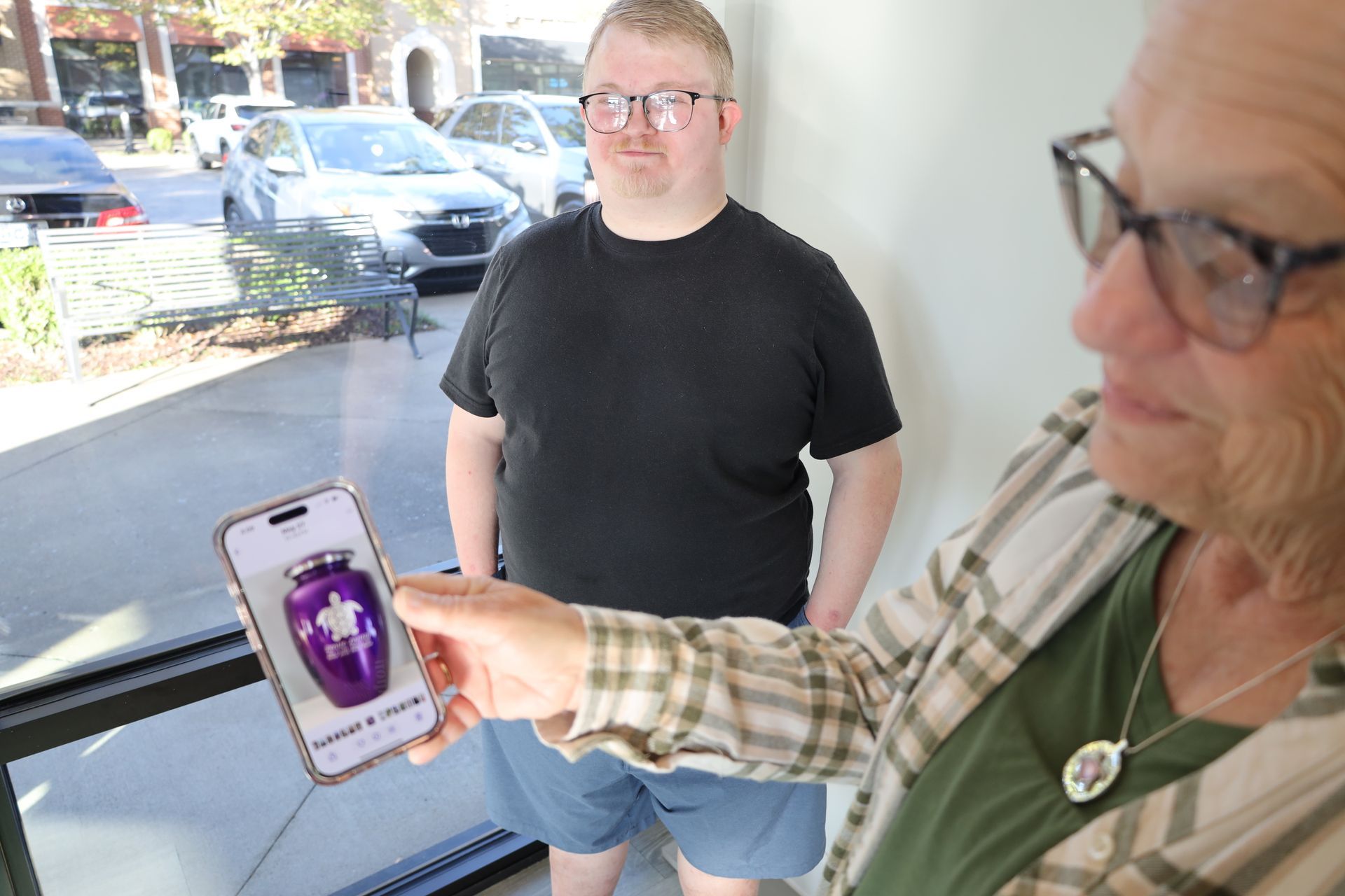 Woman holding phone, showing purple urn with floral design to a man in a black shirt. They stand near a window.