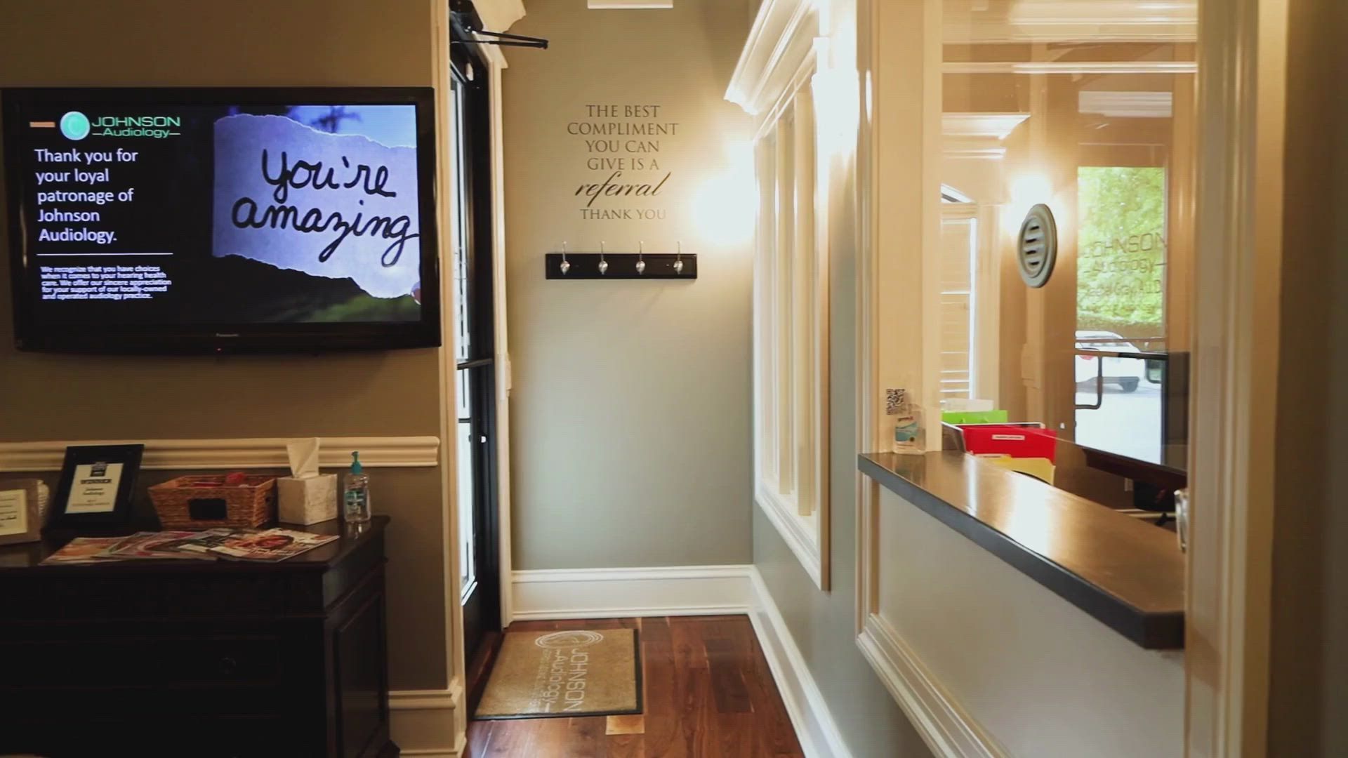 A brightly lit office hallway with a TV screen, a front desk, and inspirational wall art.