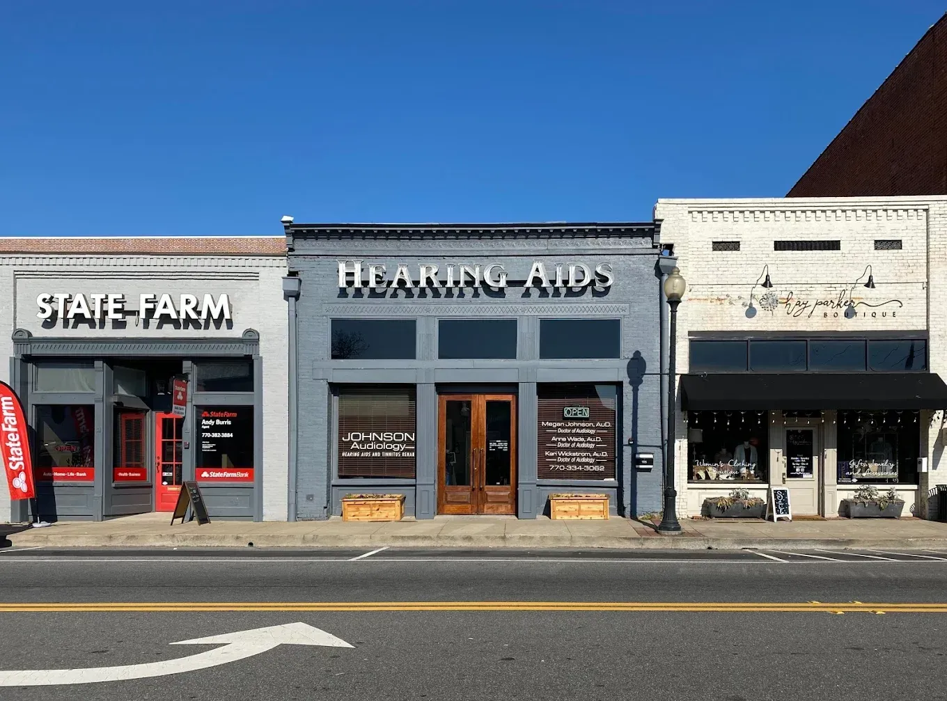 Shops on a street: State Farm, Hearing Aids, and a business under a black awning, sunny day.