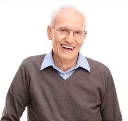 An elderly man wearing glasses and a brown sweater is smiling.