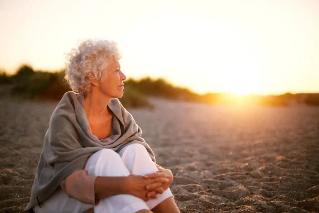 An elderly woman is sitting on the beach at sunset.