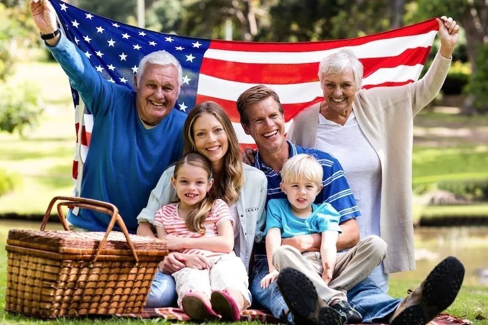 A family is sitting on a blanket in a park holding an american flag.