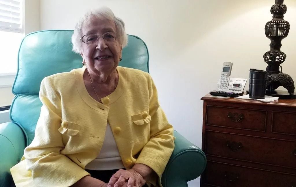 An elderly woman in a yellow jacket is sitting in a chair.