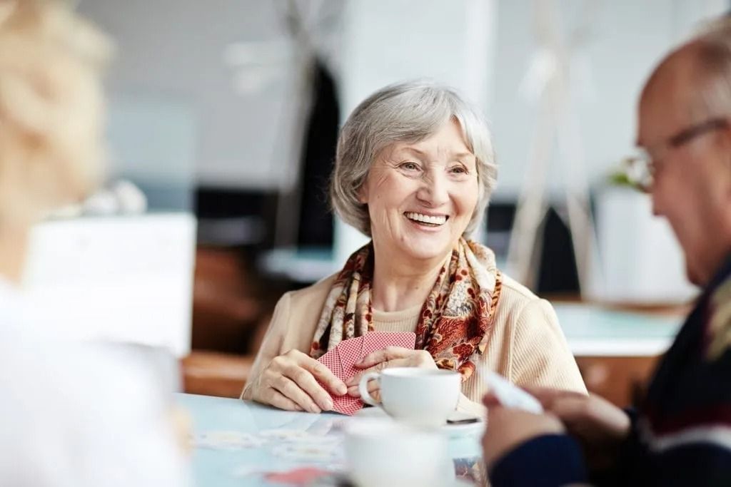 An elderly woman is sitting at a table with a cup of coffee and playing cards.