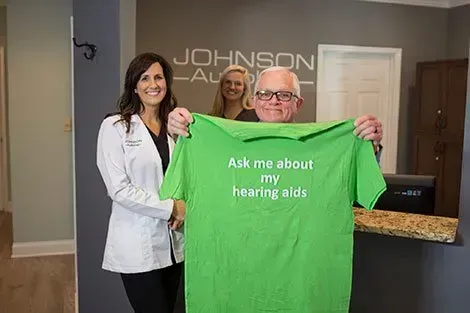 A man is holding a green t-shirt that says `` ask me about my hearing aids ''.