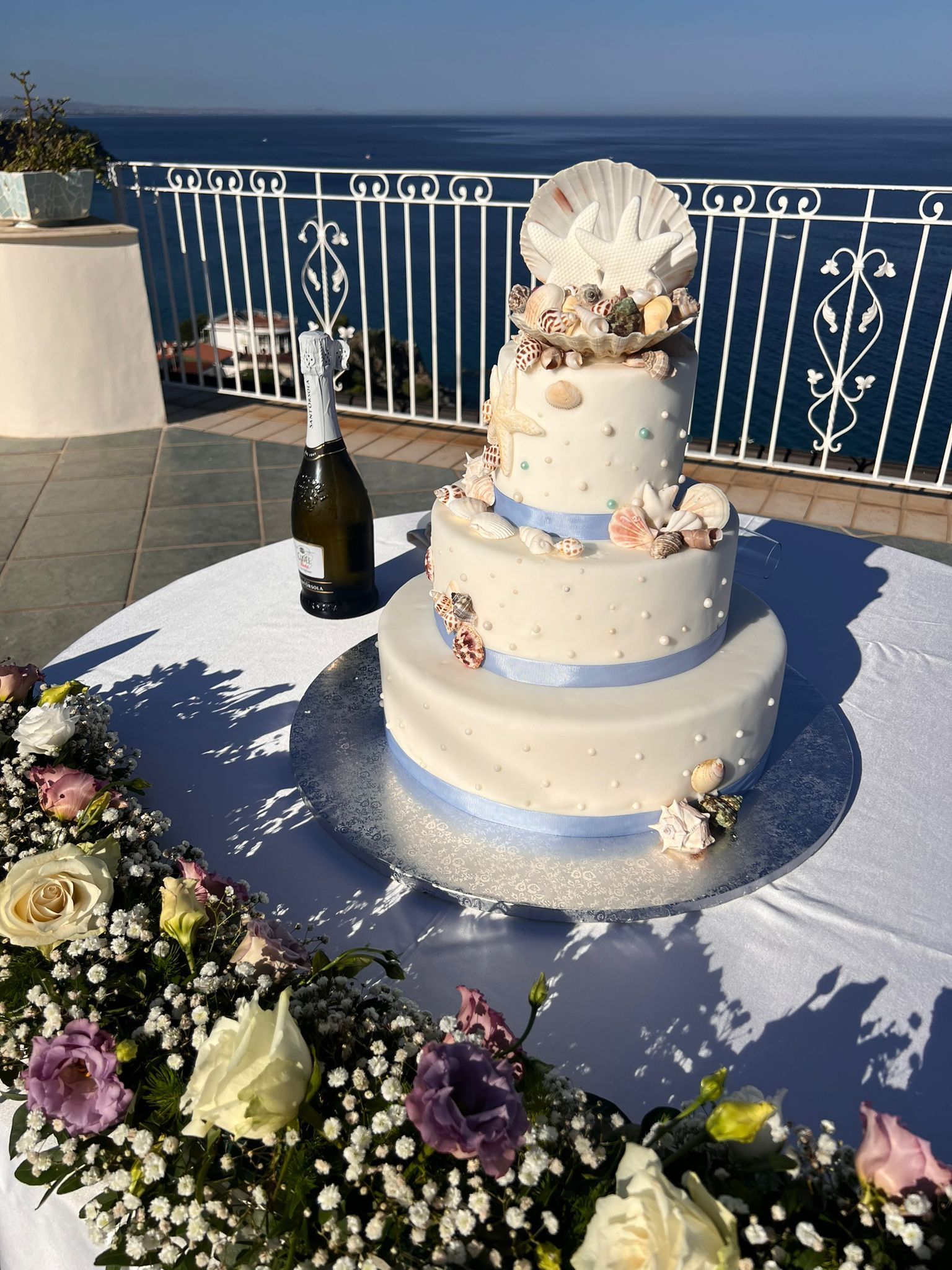 Una torta nuziale è appoggiata su un tavolo con dei fiori e una bottiglia di champagne.