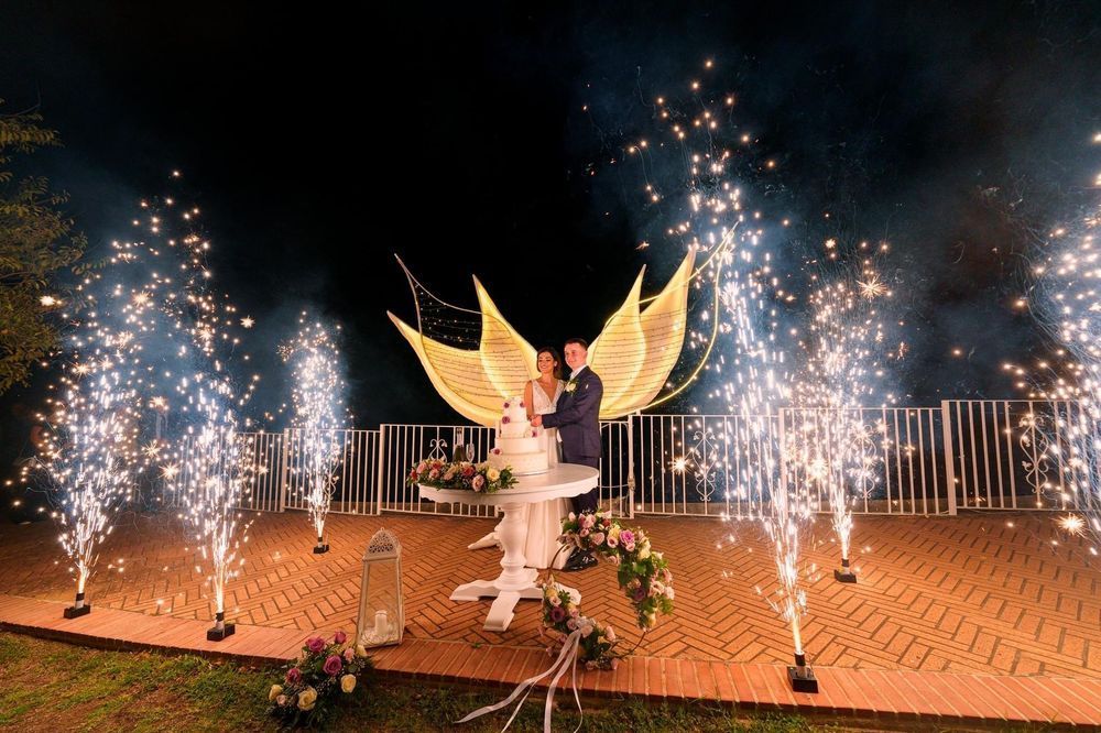 Gli sposi sono in piedi accanto a un tavolo con fuochi d'artificio alle loro spalle.