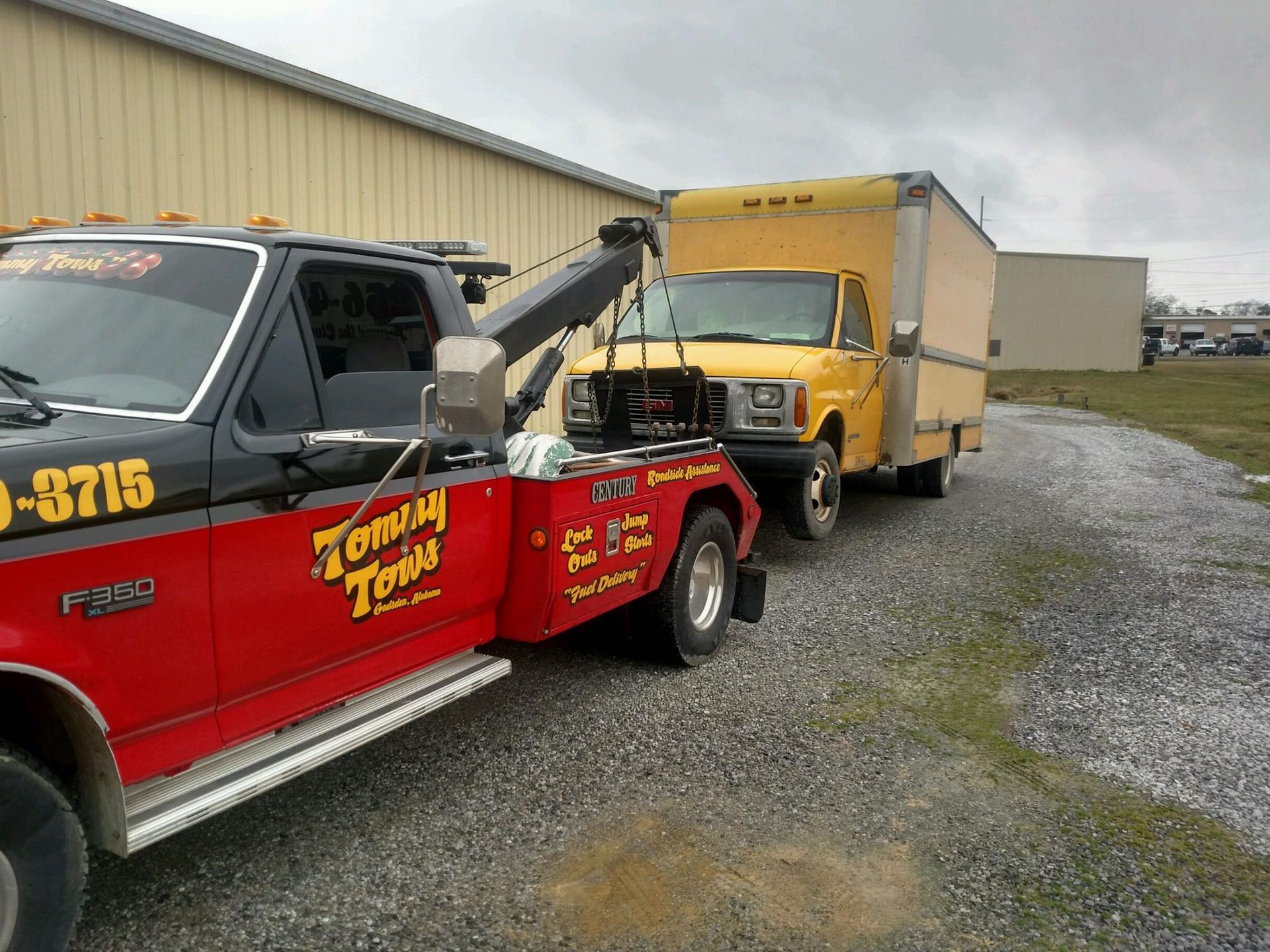 Tow truck towing a yellow box truck on a gravel lot; overcast day.