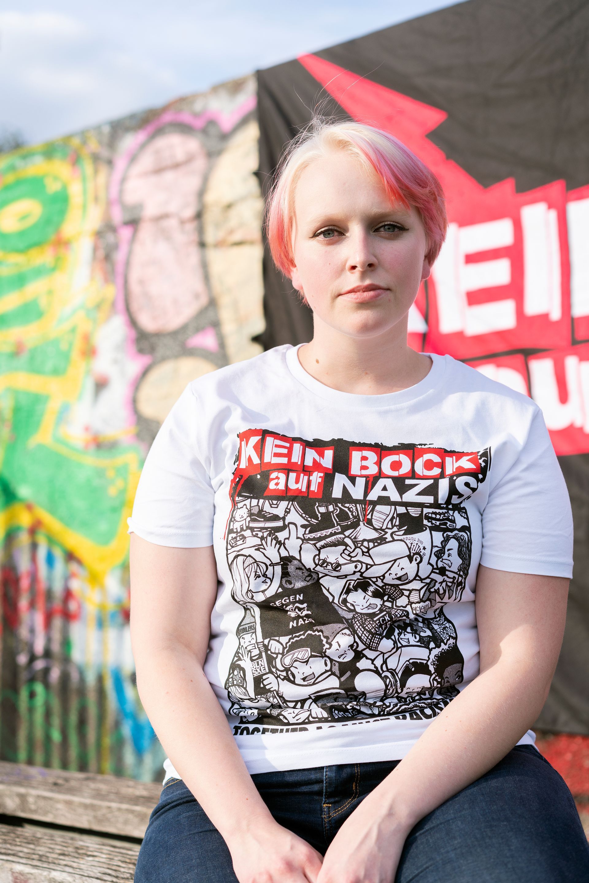 Eine Frau mit pinkfarbenen Haaren und einem T-Shirt mit der Aufschrift „No Rock for Nazis“ sitzt vor Graffiti.