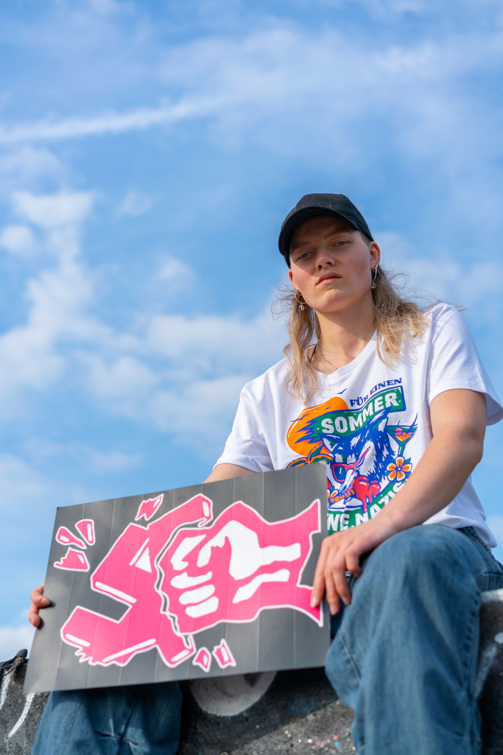 Person mit Graffiti-Schild, schwarzer Kappe und weißem Hemd, blauer Himmel im Hintergrund.