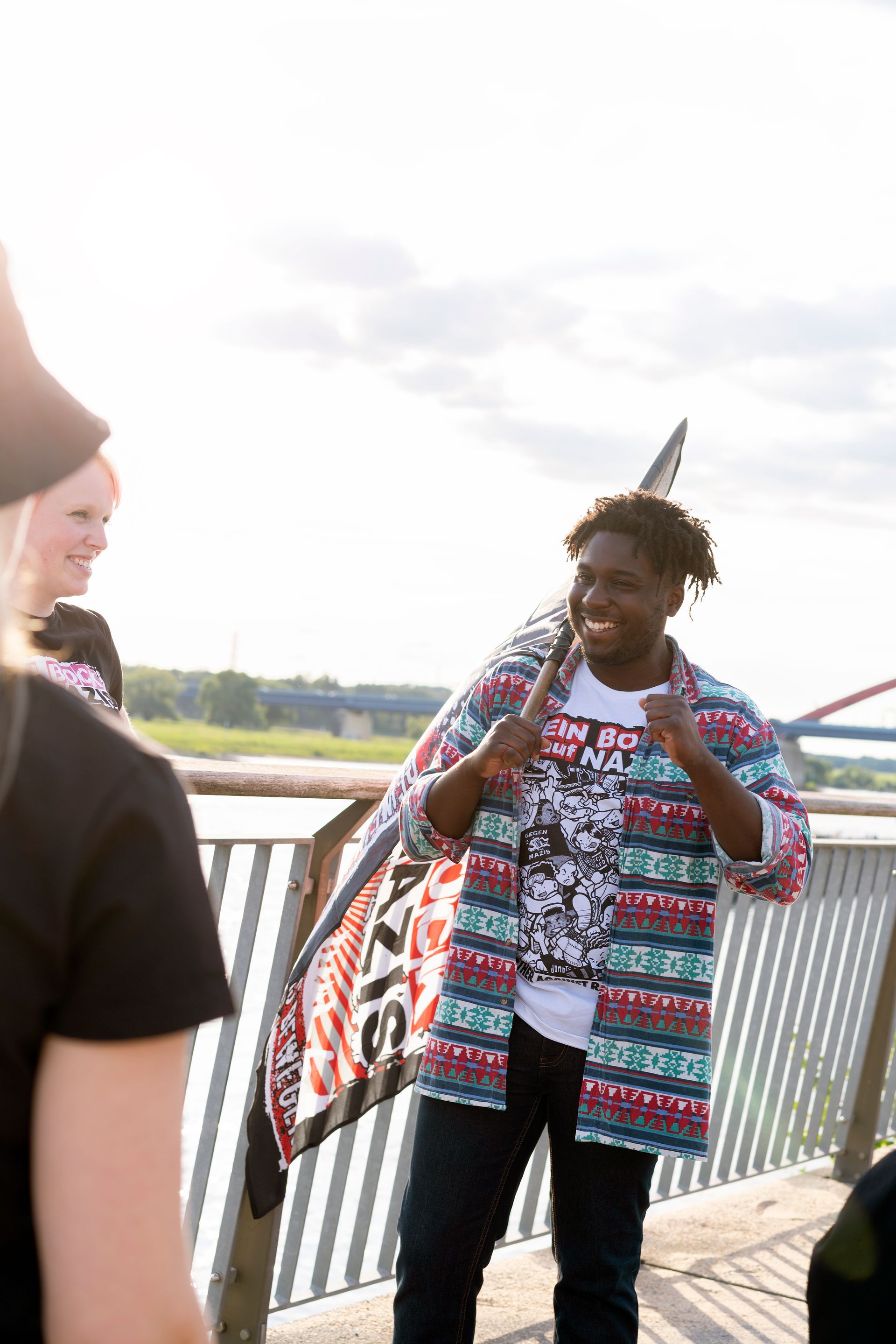 Eine lächelnde Person mit einer Fahne steht auf einer Brücke. Andere Personen befinden sich in der Nähe.
