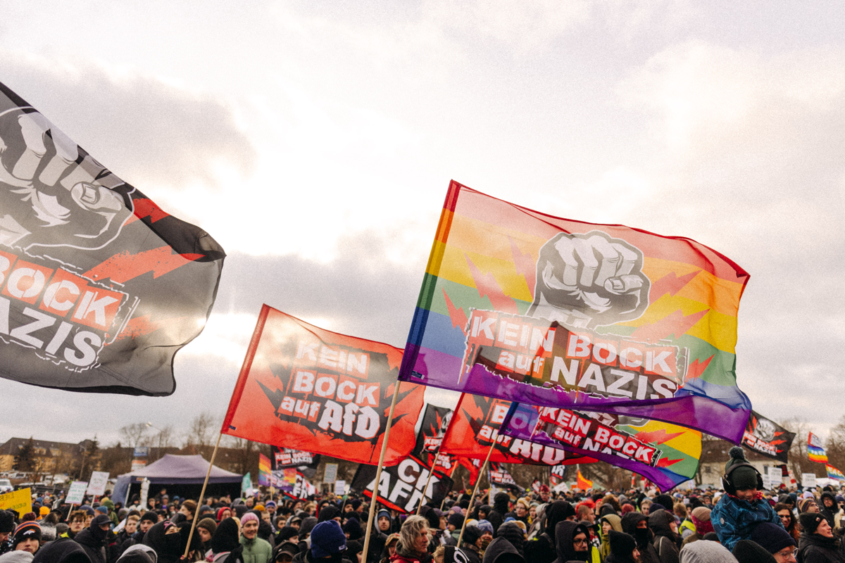 Menschen bei einer Protestkundgebung schwenken Fahnen mit Parolen gegen Nazis. Regenbogenfahne zu sehen. Bewölkter Himmel.