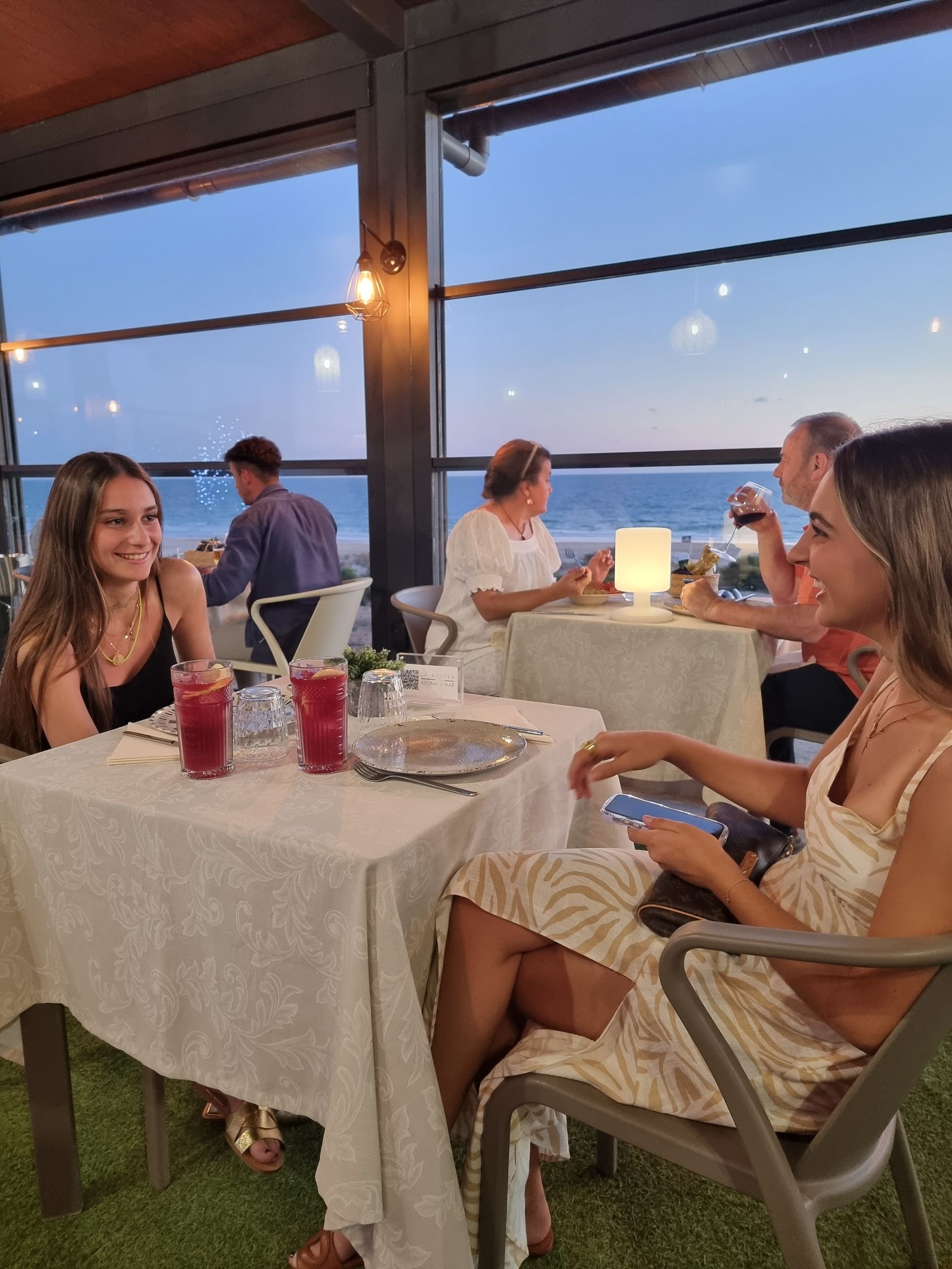 Personas cenando al aire libre junto al agua al anochecer, con sonrisas, bebidas y una lámpara encendida.