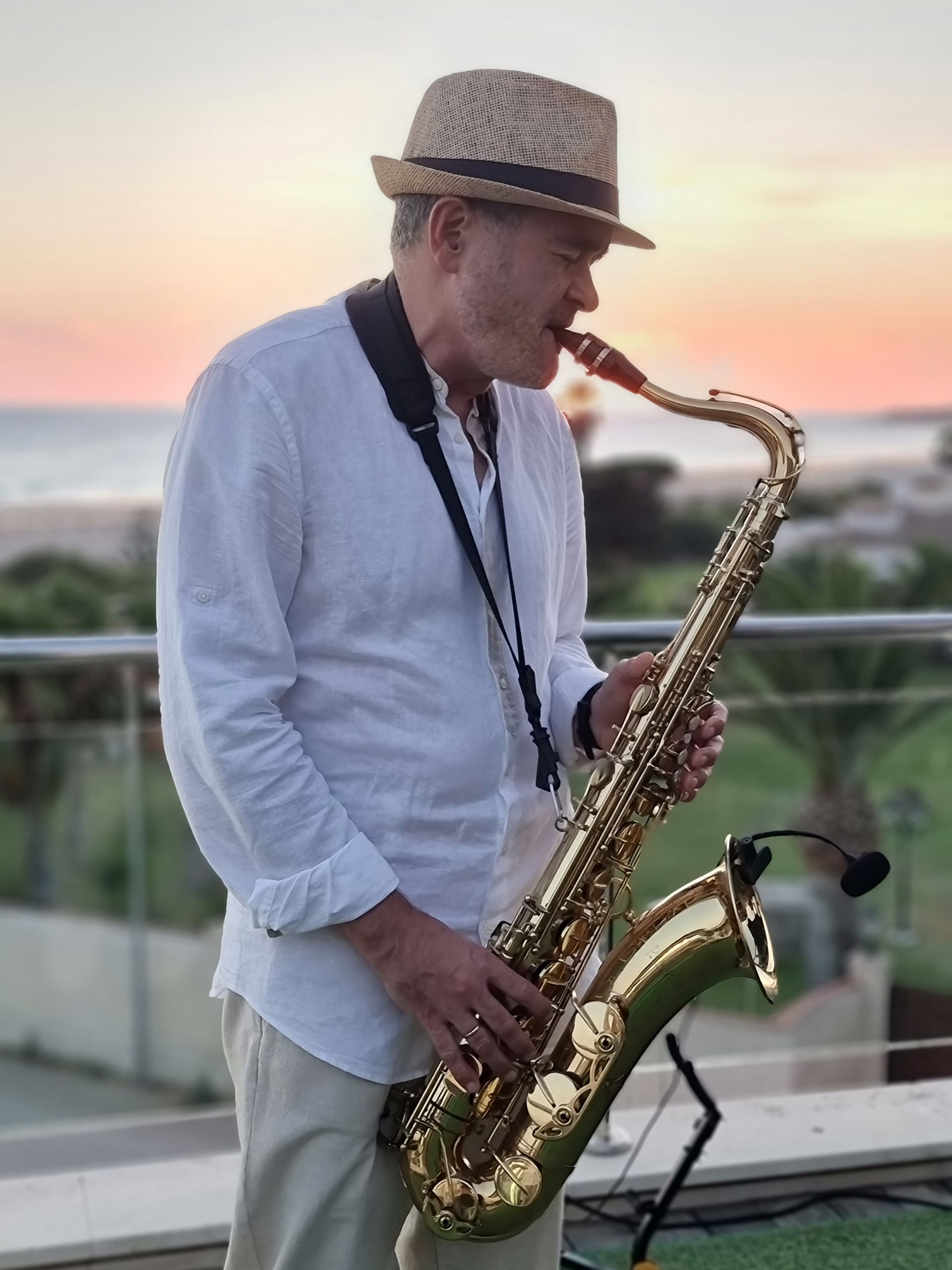 Hombre tocando el saxofón al aire libre al atardecer, vistiendo una camisa blanca y un sombrero de paja.
