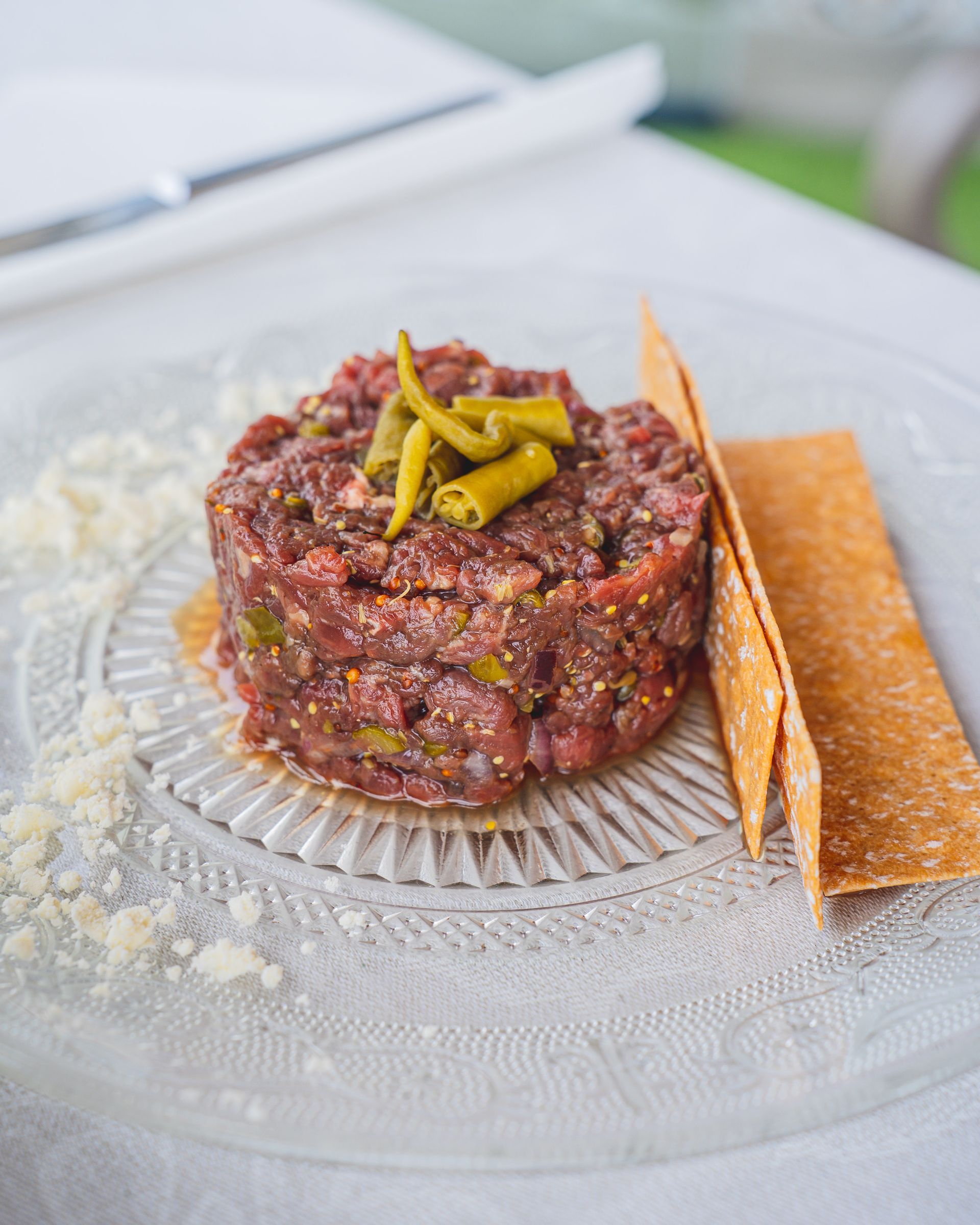 Steak tartar en un plato transparente con galletas y pimientos encurtidos, sobre un mantel blanco.