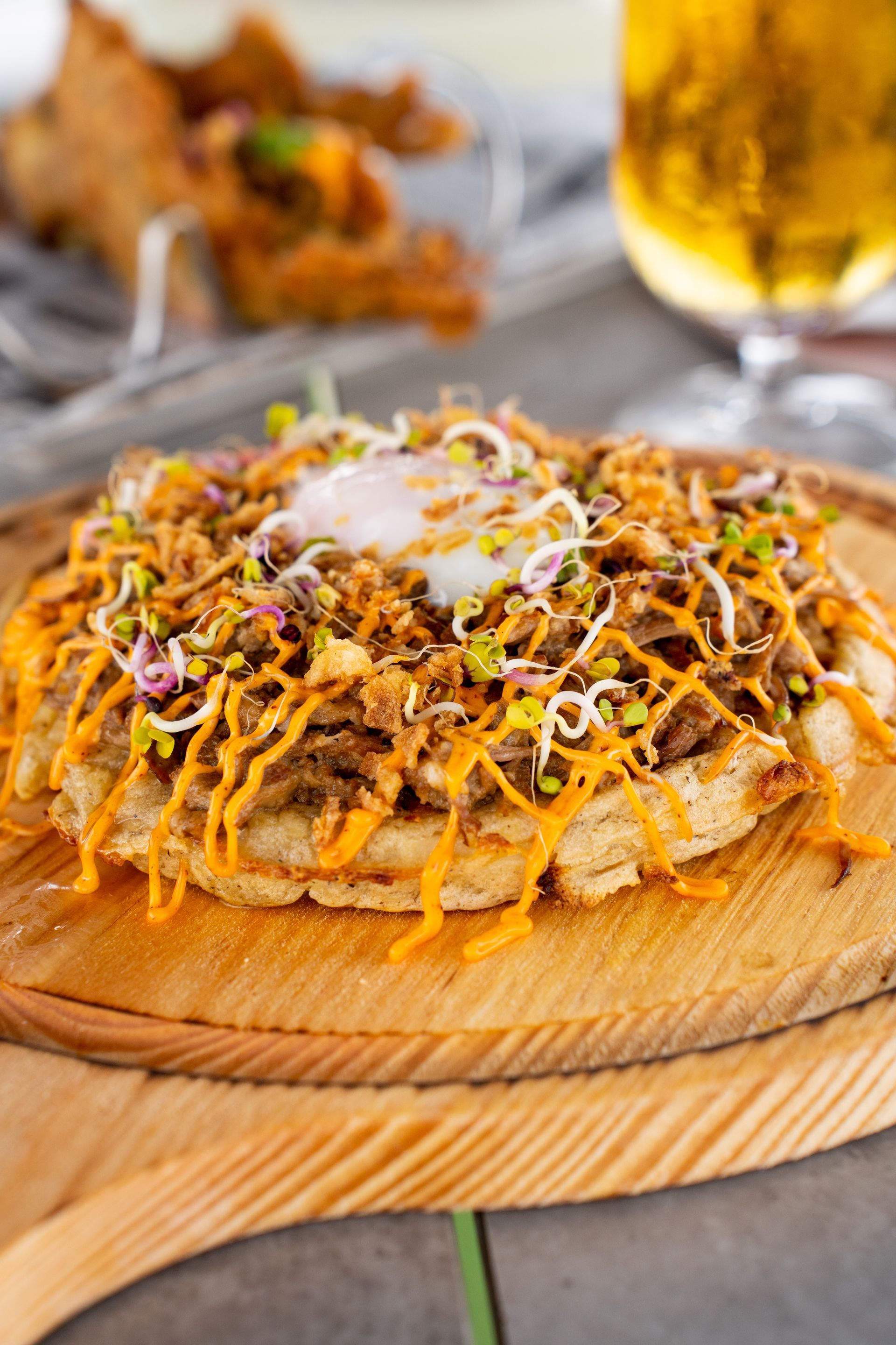 Gofre cubierto con carne desmenuzada, brotes, salsa y un huevo escalfado, sobre una tabla de servir de madera.