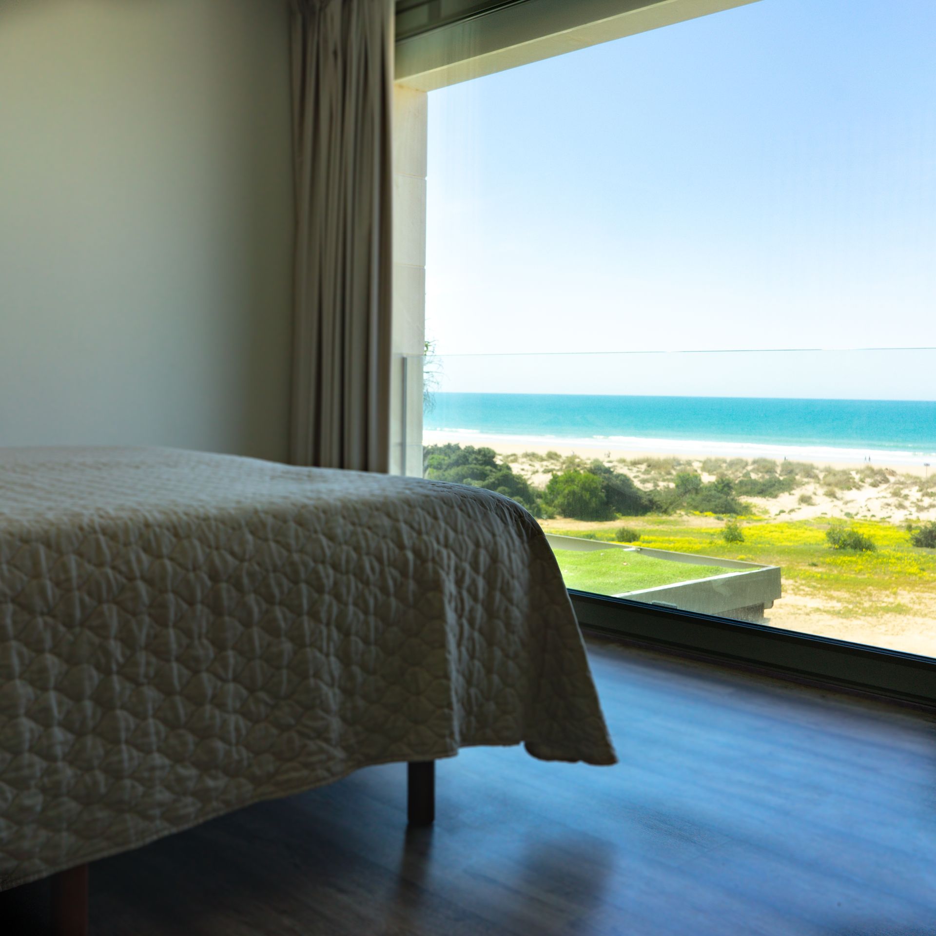 Dormitorio con cama, cortina y gran ventanal con vista a la playa y al océano azul.