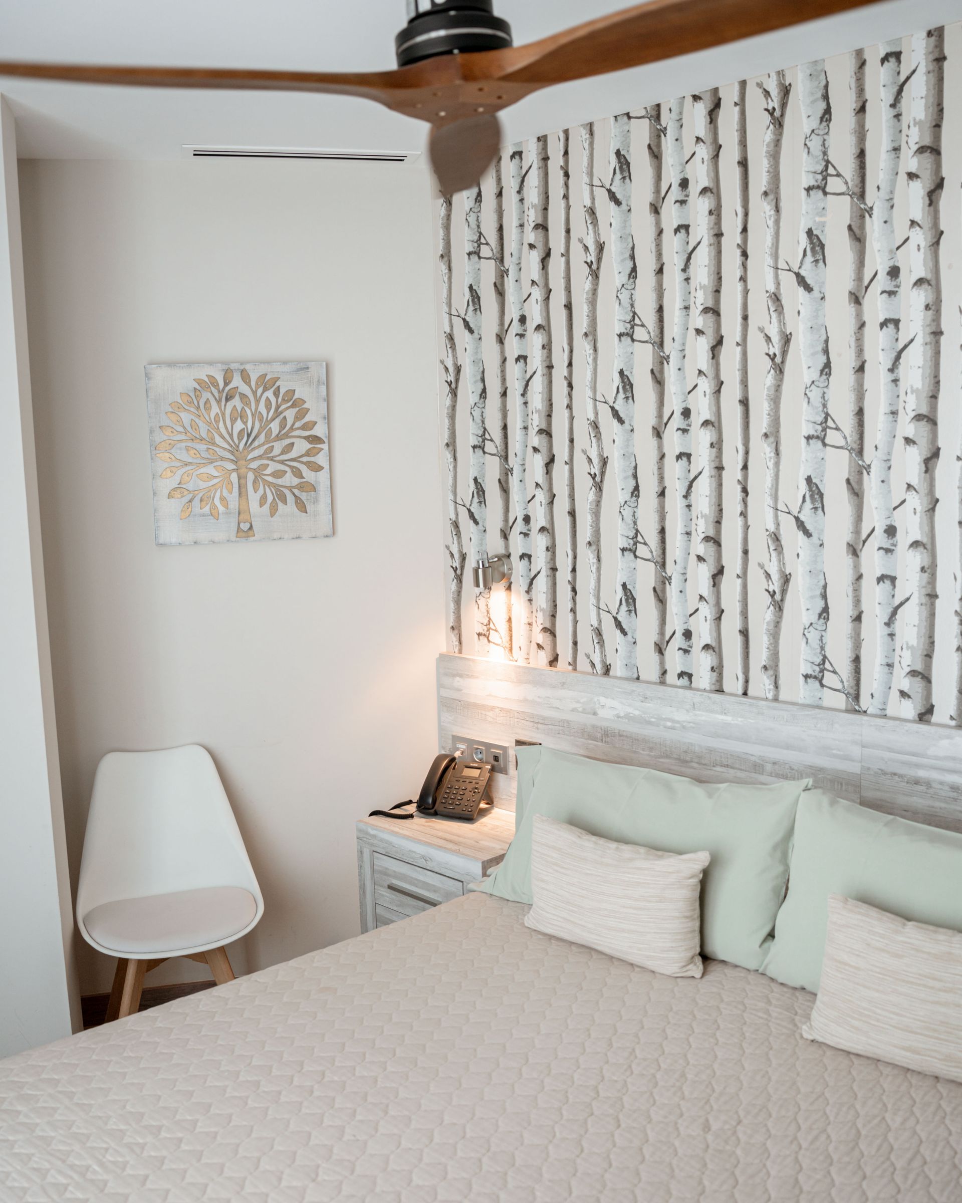 Dormitorio con pared de abedul, cama, silla y obra de arte. Colores neutros y suaves, ventilador de techo de madera.