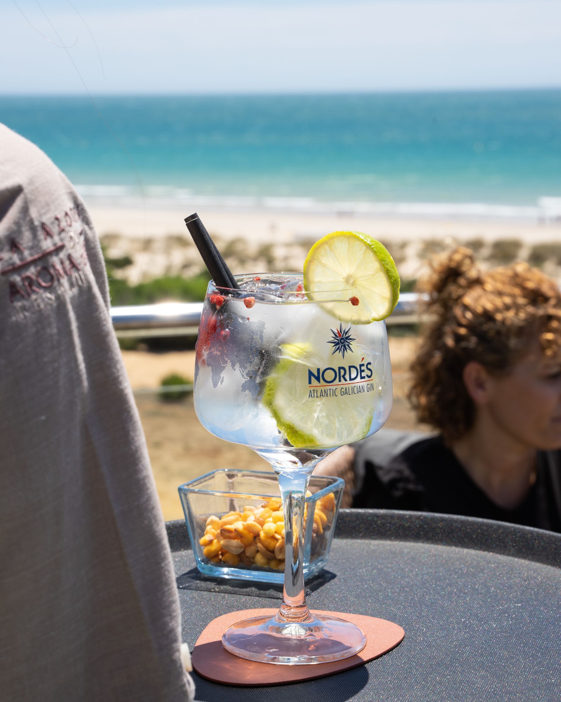 Cóctel de gin tonic con lima y frutos rojos, vista al mar de fondo.