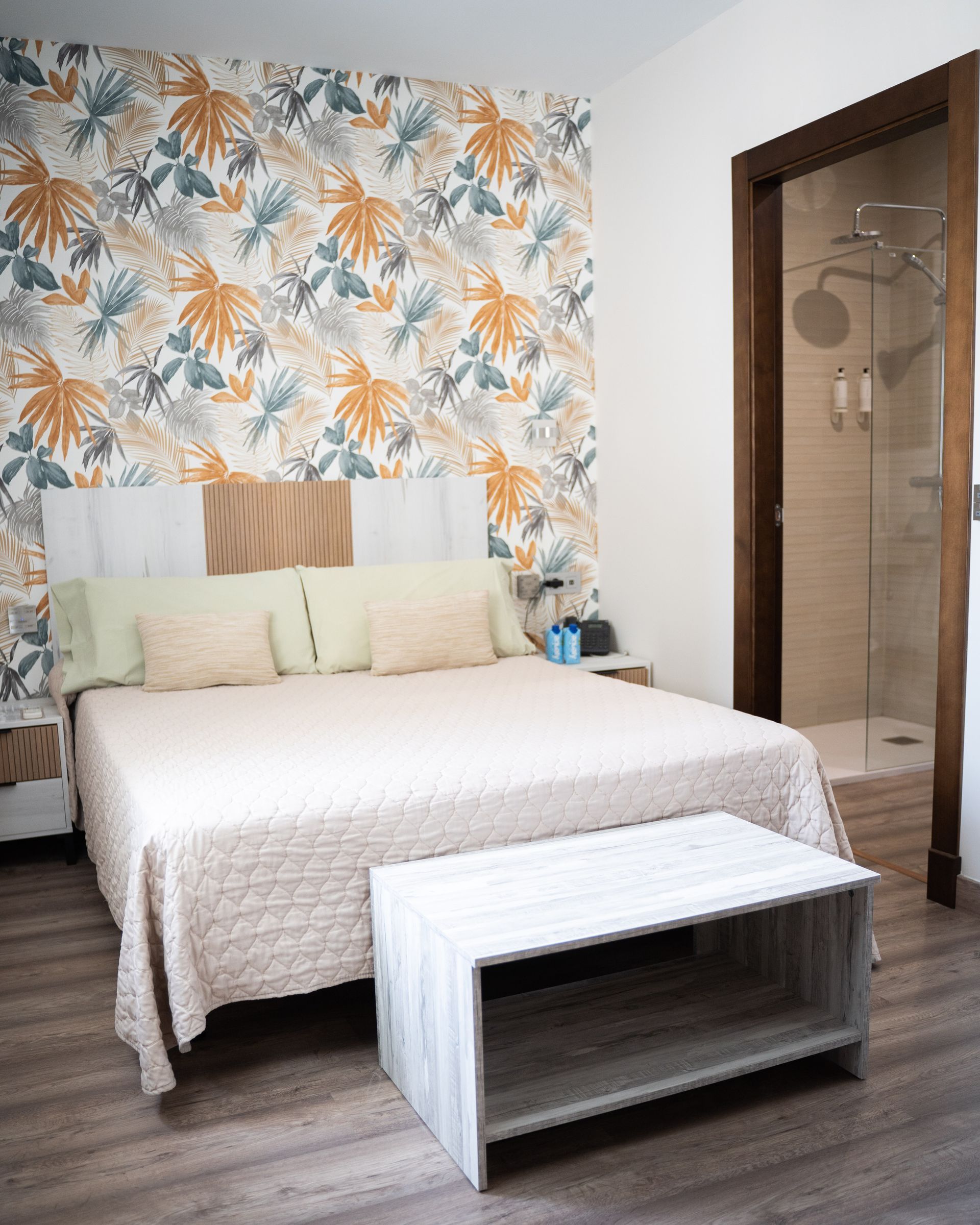 Dormitorio con papel pintado floral, cama con edredón ligero, banco y ducha visible a través de la puerta.