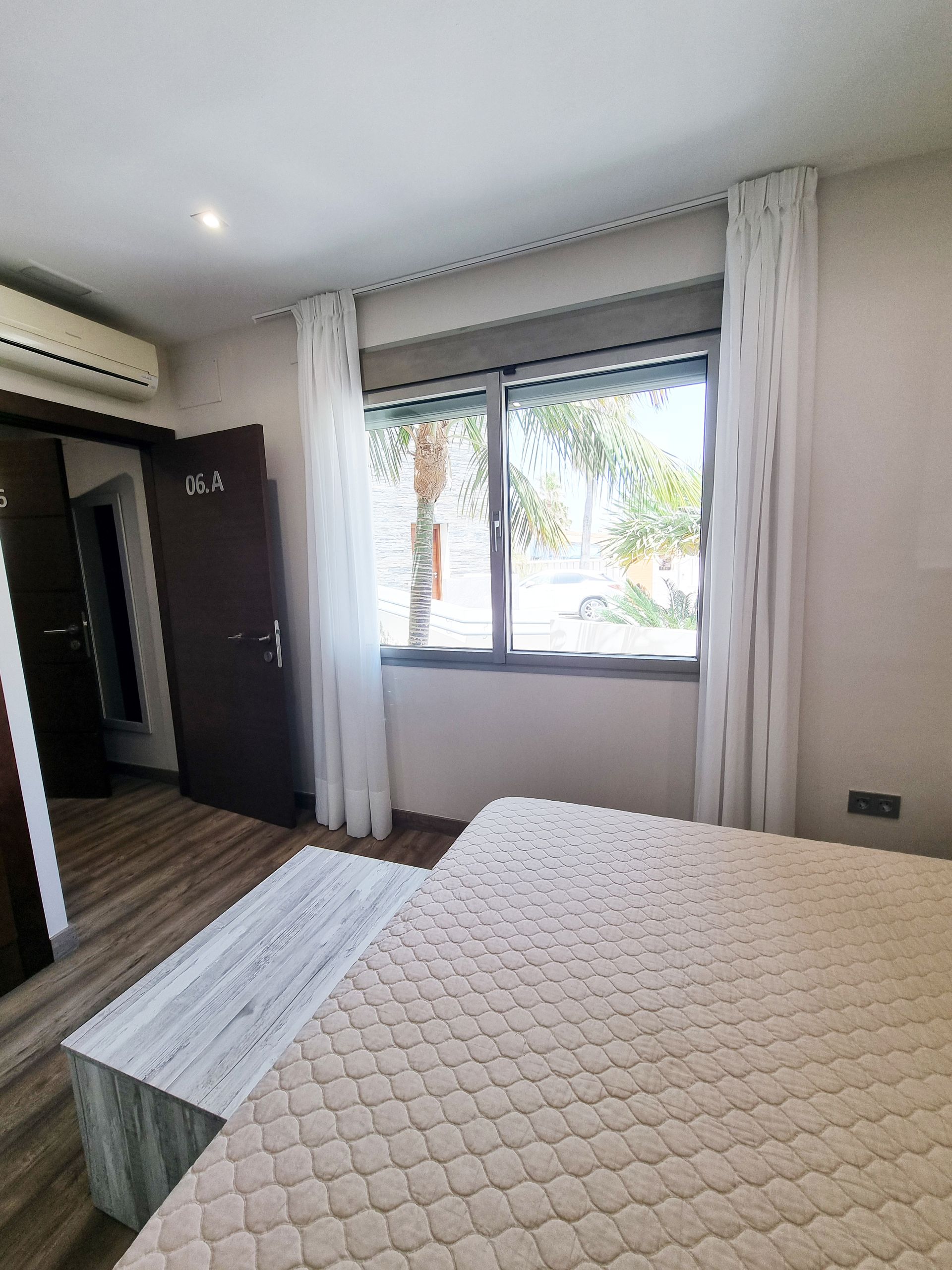 Dormitorio con cama, ventana, cortinas y un banco de madera. Los colores claros predominan en la escena.