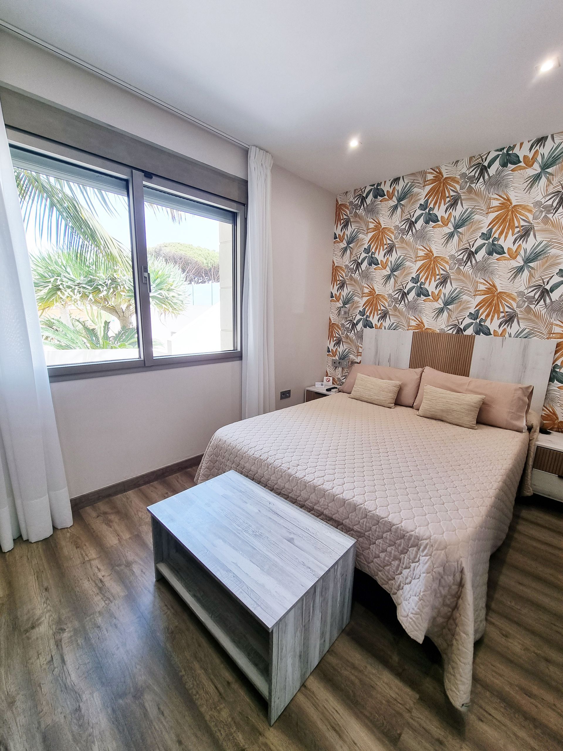 Dormitorio con cama doble, papel pintado floral, ventana y suelo de madera. Hay un banco a los pies de la cama.