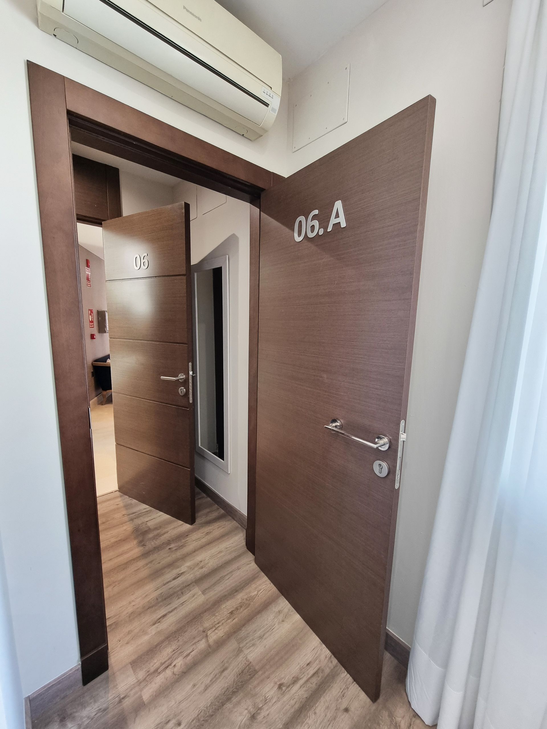 Pasillo con tres puertas marrones. Una abierta, dos cerradas. Se ve el número 06A.