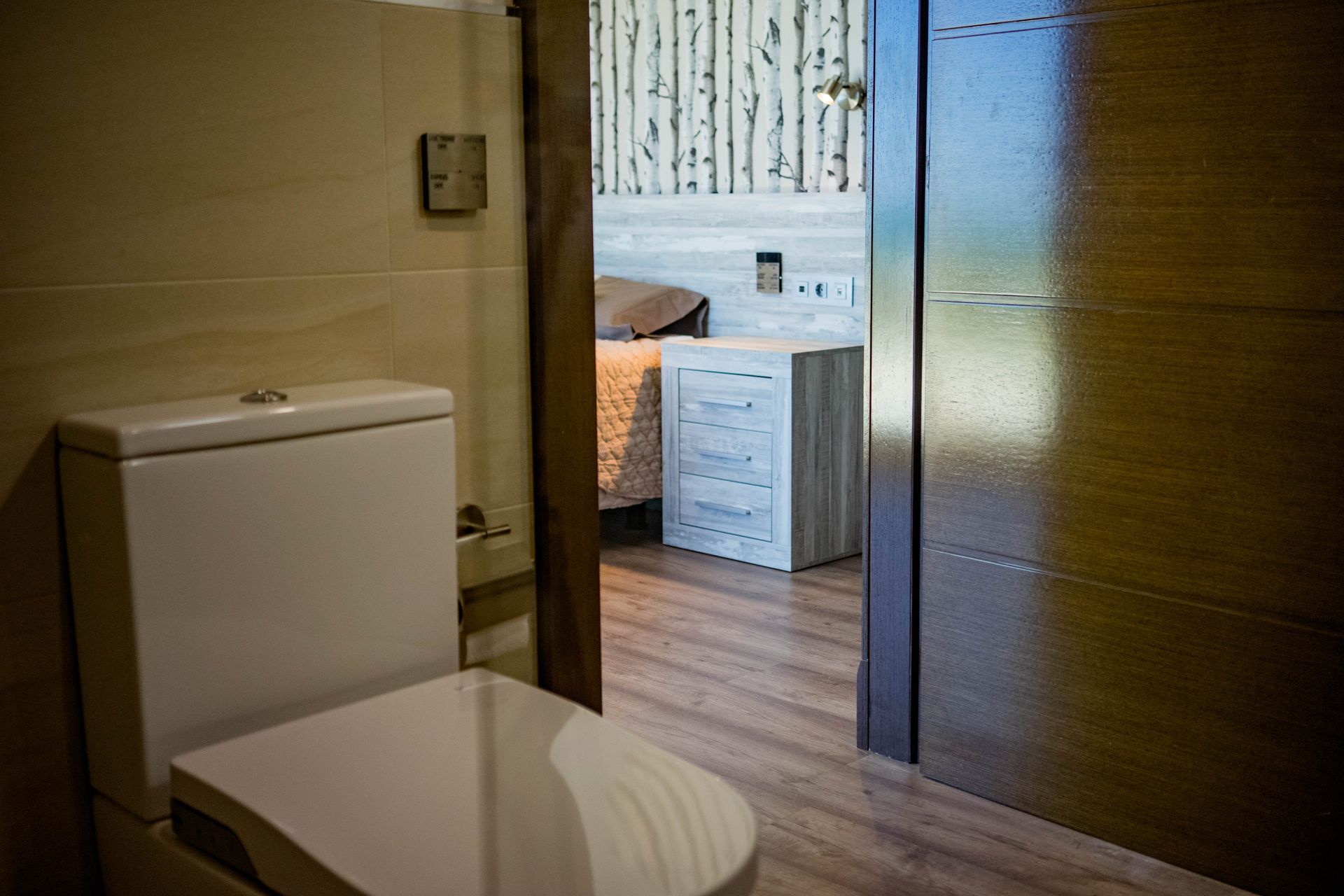 Inodoro en el baño, la puerta parcialmente abierta revela un dormitorio con cama y mesita de noche.