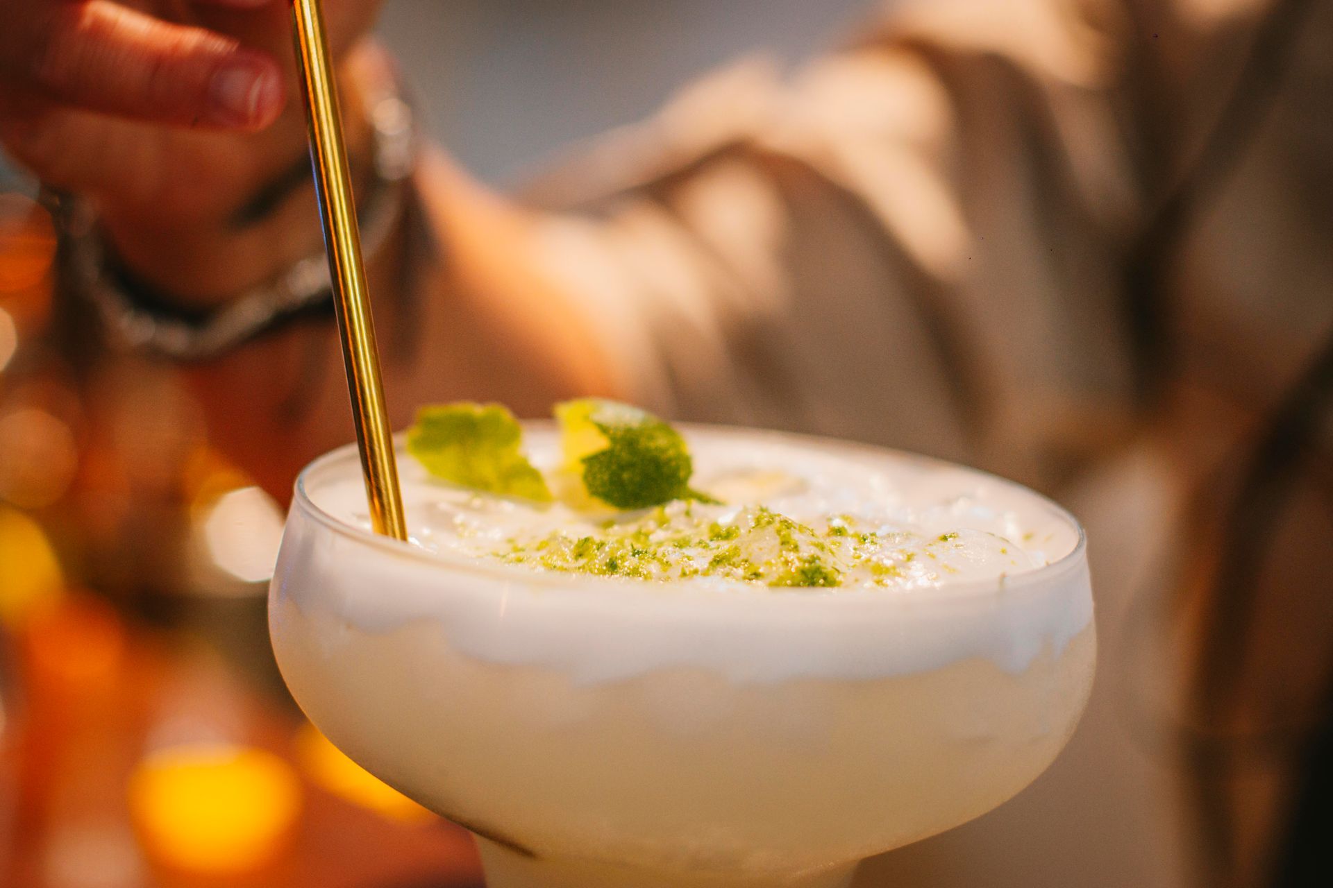 Cóctel en copa blanca revuelto con cuchara dorada y adornado con ralladura de lima.