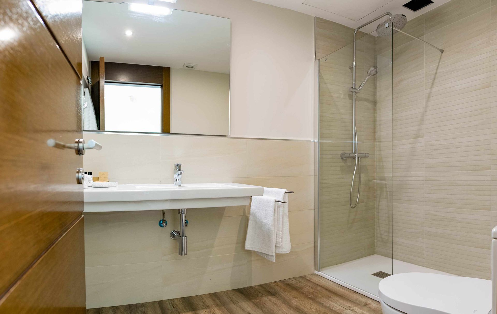 Baño con espejo grande, lavabo blanco y ducha de cristal. Puerta marrón a la izquierda.