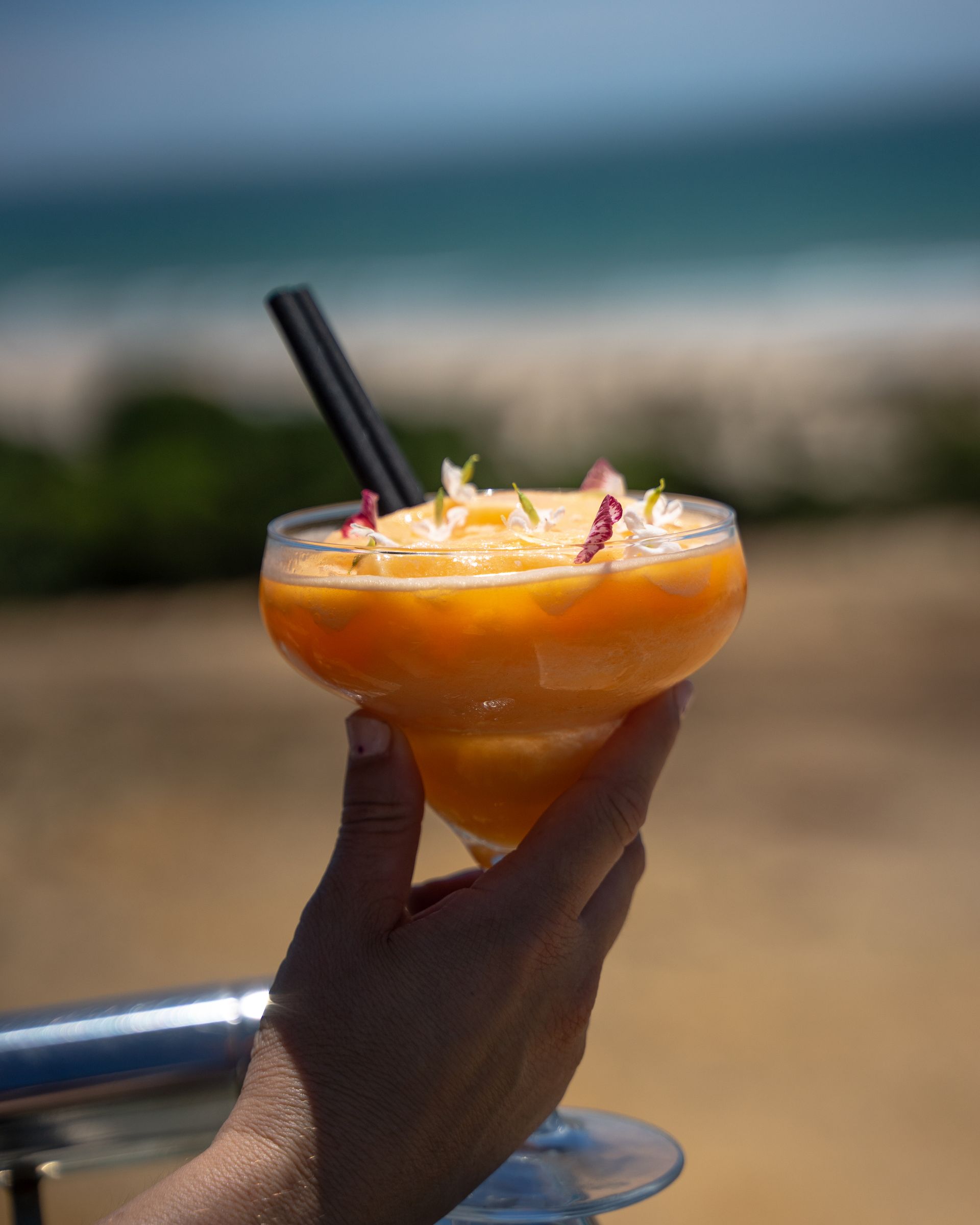 Mano sosteniendo una copa de margarita de mango con vista a la playa y al océano de fondo.