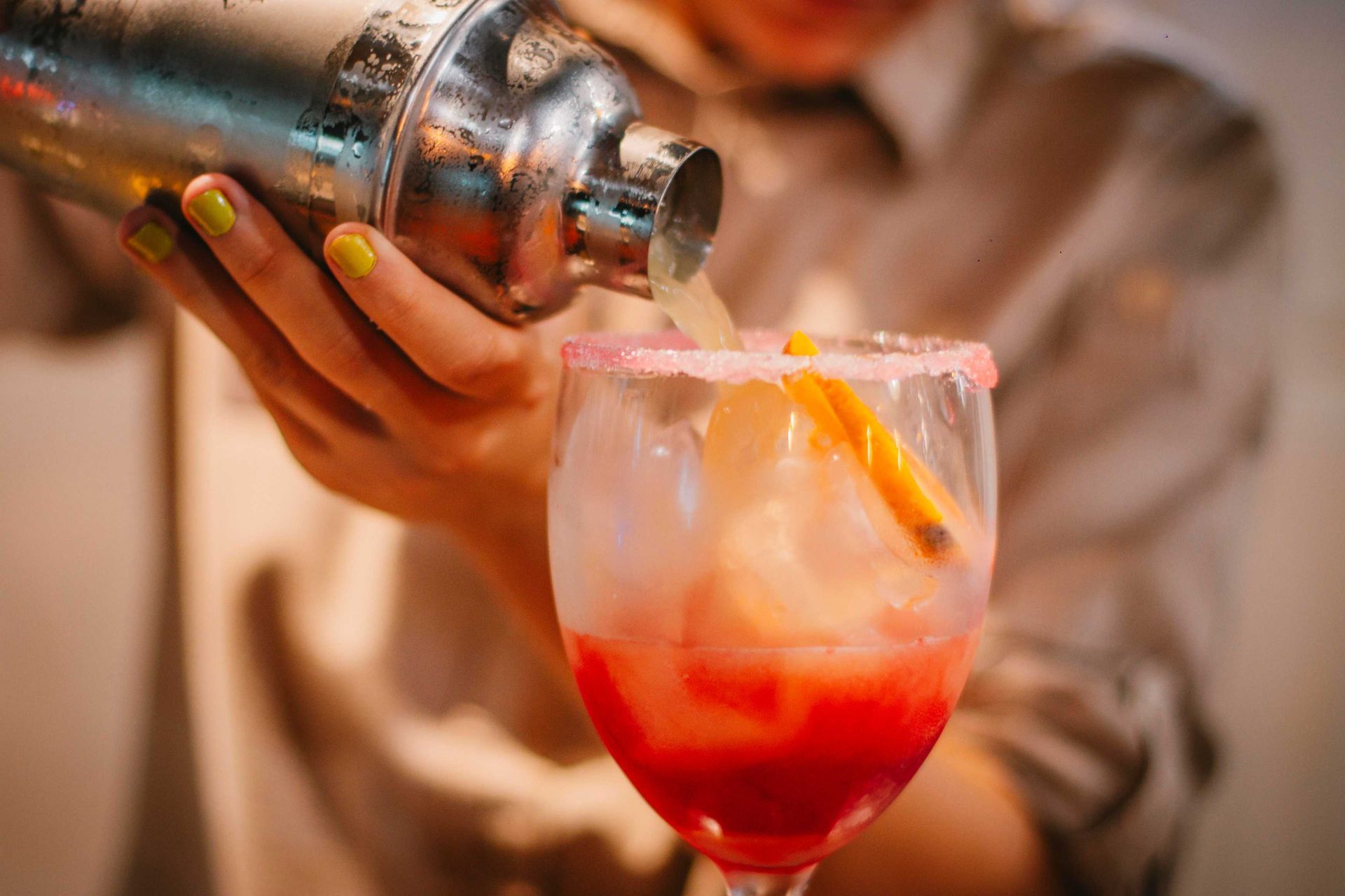 Camarero sirviendo un cóctel desde una coctelera en un vaso con borde rosa y guarnición de naranja.